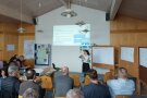 Ein Bild von einem Saal mit vielen Teilnehmern auf Stühle, vorne steht eine Frau, die etwas präsentiert.