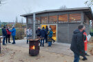 Vor einer Holzhütte stehen zahlreiche Gäste an einer Feuerstelle und unterhalten sich.