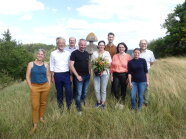 Ein Gruppenbild von mehreren Personen auf einem Feld. Eine Frau in der Mitte hält einen Blumenstrauß in der Hand. 