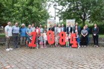 Mehrere Personen, Erwachsene und Kinder, stehen hinter großen roten Zahlen (1084).