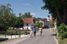 Drei Frauen laufen auf Dorfstraße mit daneben fließendem Bächlein in das Dorf. Entlang der Straße giebel- und traufseitige Wohnhäuser. Kirchturm im Hintergrund