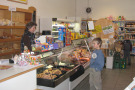 Junge wählt an der Wursttheke im Dorfladen im Dorfladen aus. Wartende Verkäuferin hinter der Theke; hinter ihr Regale mit Bäckereiprodukten.