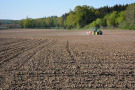 Ein Traktor ist mit einer Maissämaschine auf einem großen Acker unterwegs. Das Feld ist im Hintergrund von Grünzügen eingerahmt. 