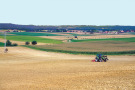 Landschaft mit Traktor bei der Feldbestellung und Dorf im Hintergrund.